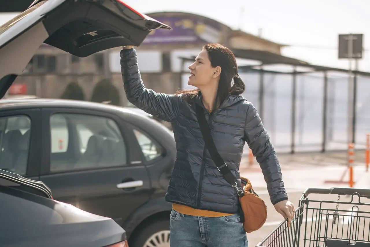 Young woman closing car trunk and holding shopping cart on parking
