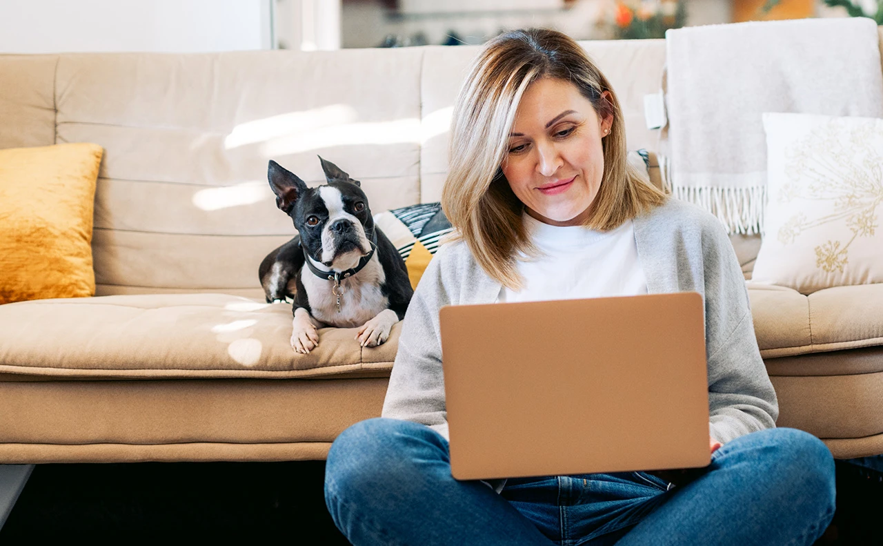 A middle age woman sitting on the floor looking at a computer. Her dog is sitting behind her on a couch.
