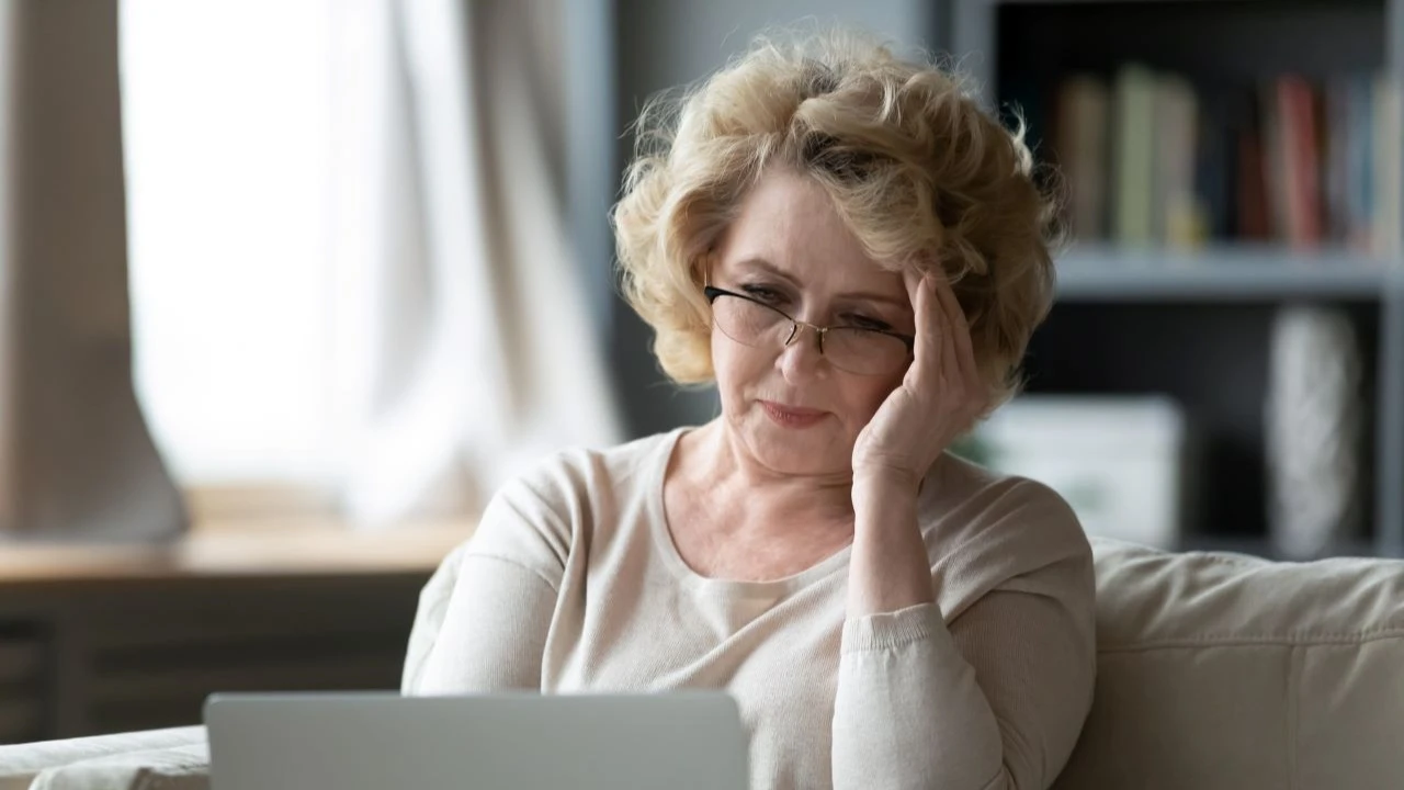 A mature woman sitting on a couch using a laptop feels stressed and needs assistance with debt