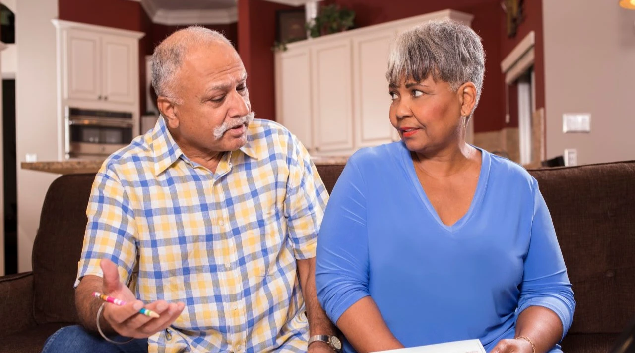 Indian man and African descent woman, senior adult couple work together to pay their monthly bills. They are calculating expenses versus budget income. Many invoices on living room table. Kitchen background.
