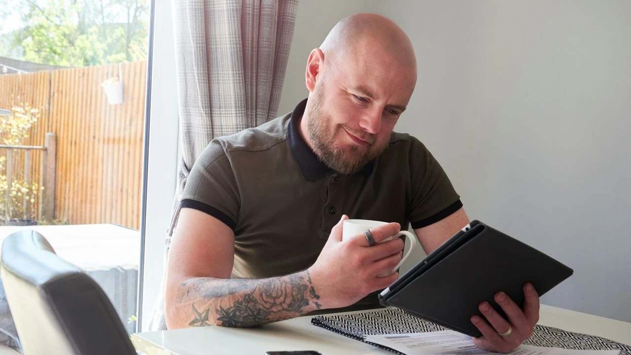 A man sits at the dining table reading on a digital tablet looking at his credit score