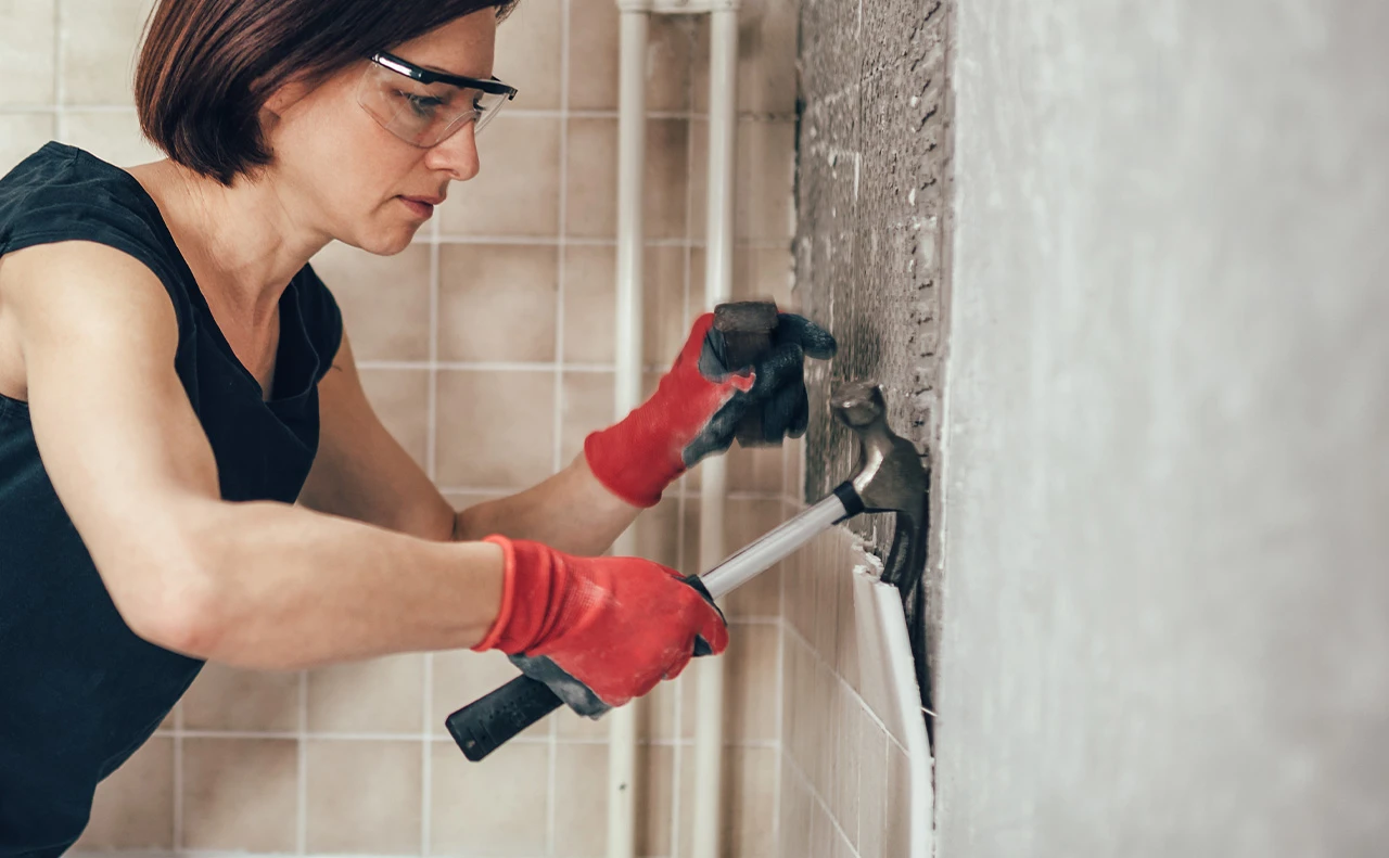 A middle age woman wearing a sleeveless black shirt, goggles, and gloves, using a hammer to do home remodeling and tearing out tiles