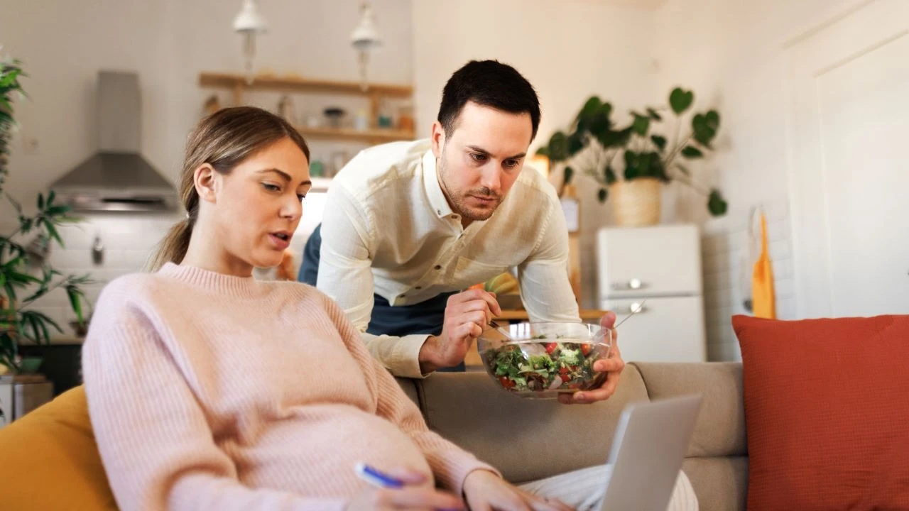 Young casually clothed man bringing salad to his working pregnant wife, she is sitting on the sofa and using laptop