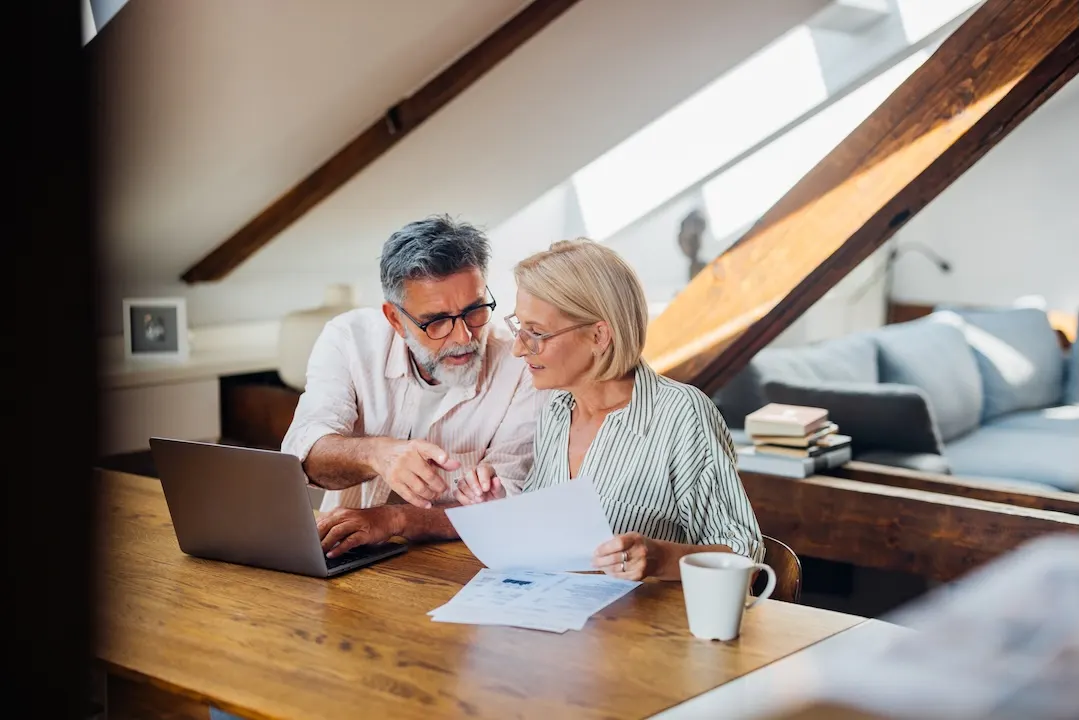 Older couple reviews paperwork at a laptop.