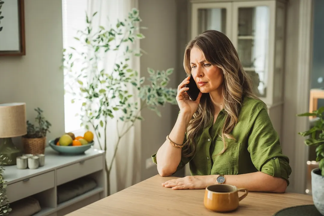 Upset woman talks on the phone while sitting at her dining table.