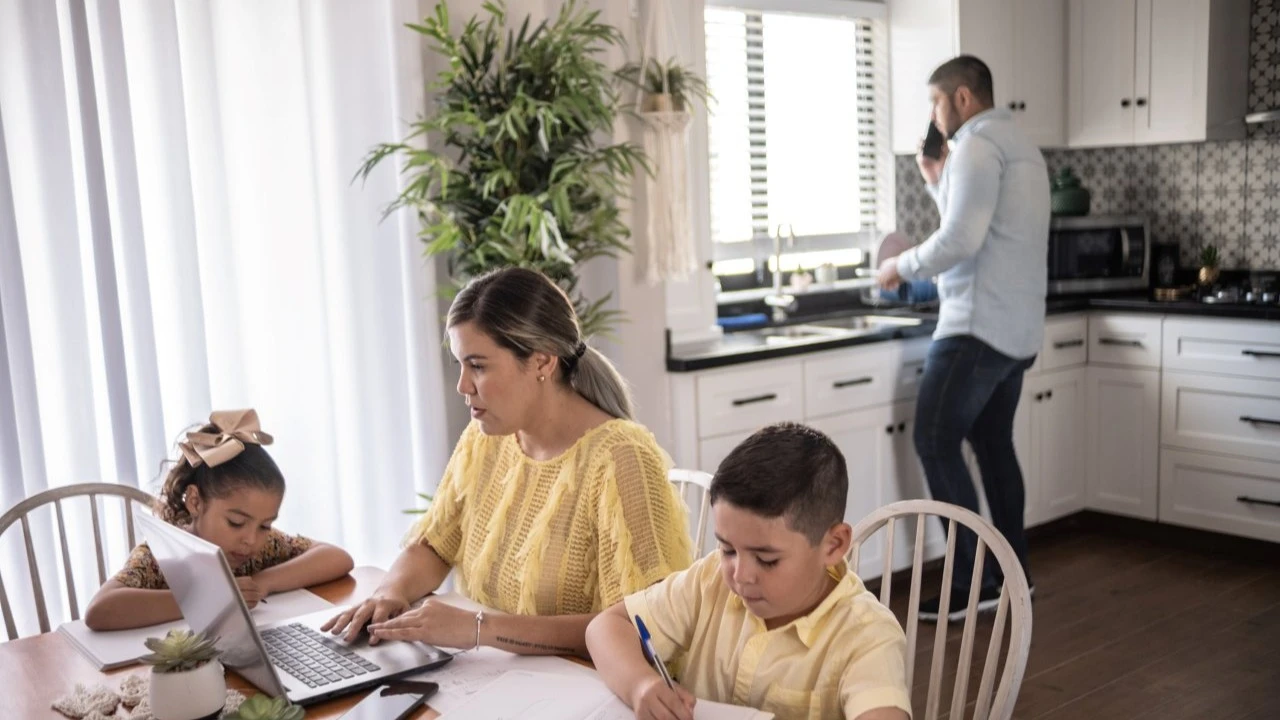  mother working and children studying at home in kitchen. Father on phone in the background