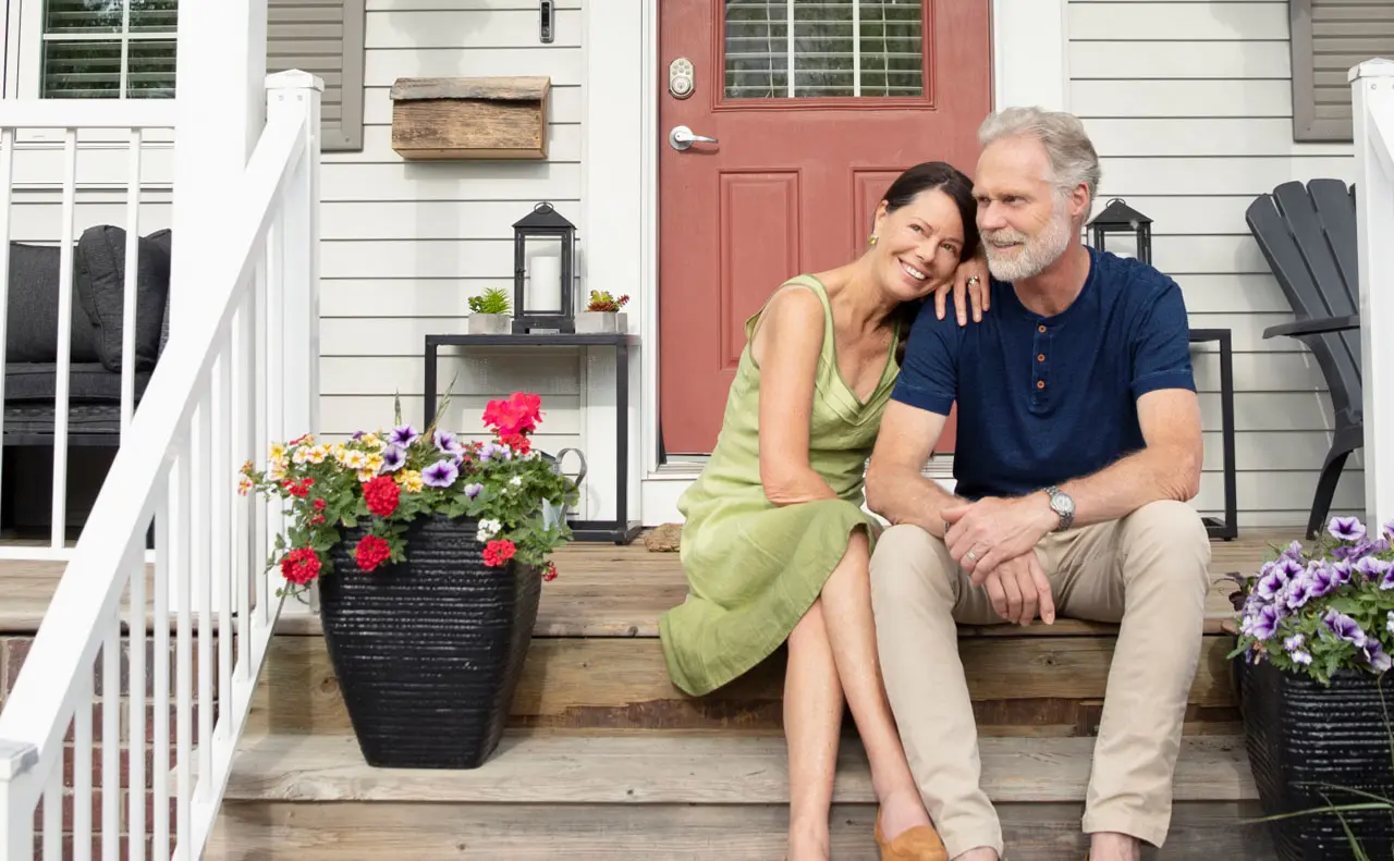 A middle age man and woman sitting on a steps near the front door with the woman resting on his shoulder and flowers on each side of them