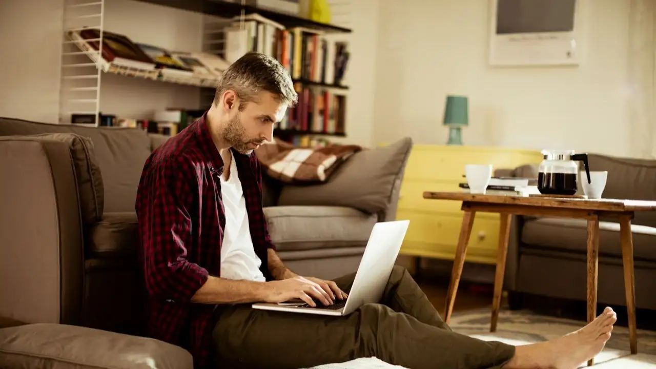Young man sitting on floor in living room with papers and computer trying to figure out finances.